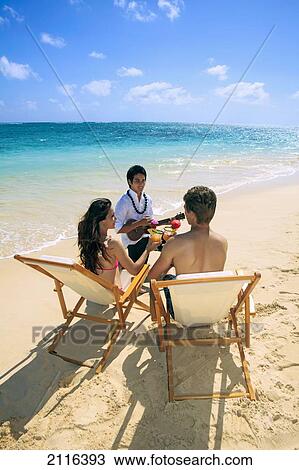 Stock Image - Hawaii, polynesian man plays ukulele for a young couple on the beach.. Fotosearch