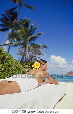 Stock Photo - Hawaii, Woman Lay On Table Along Beach, Tropical Scenery Topless With Towel Yellow Hibiscus Flower In Hair Back Angle. Fotosearch