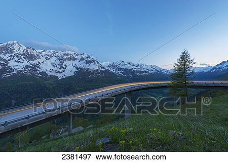 Stock Photo - Headlights blurred on a road with the swiss alps in the background; San Gottardo, Ticino, Switzerland. Fotosearch