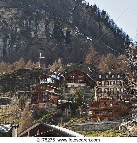 Stock Photograph - Houses On The Side Of A Hill With A White Cross On A High Rock Ledge; Zermatt Valais Switzerland. Fotosearch