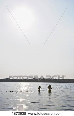 Stock Photography - Huatulco, Mexico; Two People Standing In The Water With The Sunlight Reflected On The Water. Fotosearch