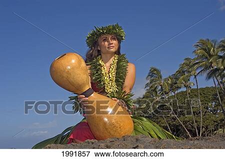 Stock Photo - Hula Dancer With Haku Lei In Ti-Leaf Skirt On Rocky Shore Holding Ipu, Palm Tree Background. Fotosearch