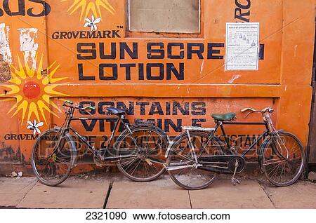 Stock Image - India, Karnataka, Two Bikes Leaning Against Wall Advertising Sun Screen; Gokarna. Fotosearch