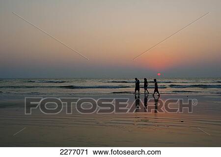 Stock Image - Indian family walking on beach at dusk; Goa, India. Fotosearch