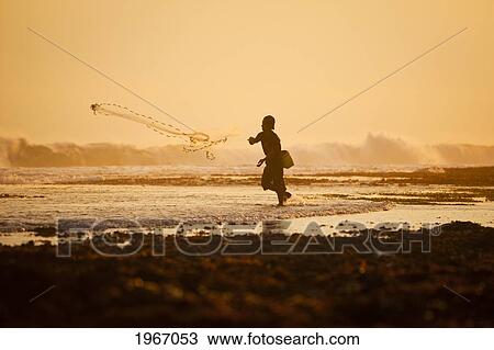 Stock Image - Indonesia, Bali, Uluwatu, Fisherman Throwing Nets At Sunset, Waves Crashing In Background. Fotosearch