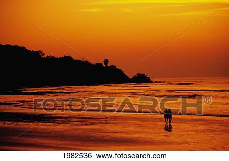 Stock Photograph - Indonesia, Jimbaran Bay, couple walking shoreline at sunset, orange sky and reflections. Fotosearch