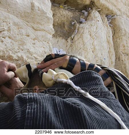 Stock Photograph - Israel. Jerusalem Old City. Western Wall. Close Up Of A Man With His Head Leaning Against The Wall And Covered With Prayer Shawl While Being Blessed By A Hand With Phylactery. Fotosearch