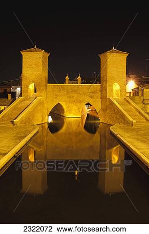 Stock Image - Italy, Emilia-Romagna, Comacchio, Stone bridge with towers and steps reflecting in water, night. Fotosearch