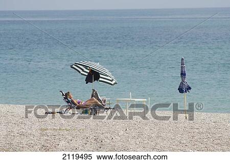 Stock Photography - Italy, Marche, Woman sunbathing on lounge chair on pebble beach with sun umbrella; Lido Tre Archi. Fotosearch