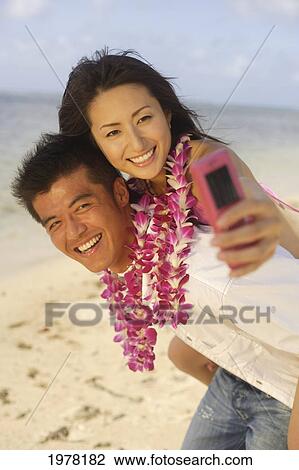 Stock Image - Japanese Couple On The Beach Wearing Leis Takes Photo Of Themselves With Camera Phone.. Fotosearch