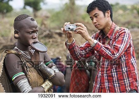 Japanese Tourist Taking A Picture Of A Slightly Annoyed Mursi Tribal Woman Wearing A Large Pottery Lip-Plate (Dhebi); Maridungka Village / Mursiland / South Omo / Southern Nations, Nationalities & People's Region, Ethiopia View Large Photo Image Stock Image - Japanese Tourist Taking A Picture Of A Slightly Annoyed Mursi Tribal Woman Wearing A Large Pottery Lip-Plate (Dhebi); Maridungka Village / Mursiland / South Omo / Southern Nations, Nationalities & People's Region, Ethiopia. Fotosearch