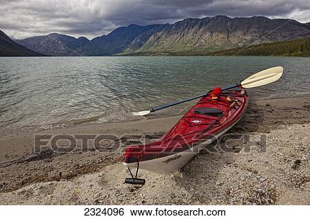 Stock Photograph - Kayak sitting on the shoreline of Kusawa Lake with the setting sunlight hitting the mountains; Yukon, Canada. Fotosearch
