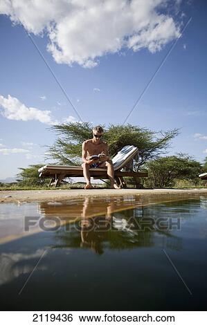 Stock Photograph - Kenya, Guest relaxing reading book beside swimming pool at luxurious Joy's Camp; Shaba National Reserve. Fotosearch
