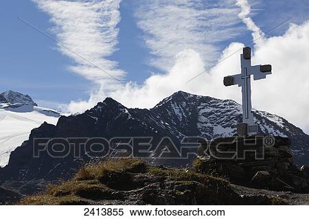 Stock Photography - Large cross on mountain peak with snow covered mountains in the background with clouds and blue sky; Hintertux, Austria. Fotosearch