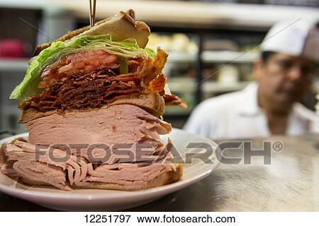 Stock Photo - Large meat sandwich with chef in the background; New York City, New York, United States of America. Fotosearch
