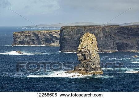 Stock Photograph - Large sea stack rock structure along rugged dramatic cliff shoreline with waves and blue sky; Kilkee, County Clare, Ireland. Fotosearch