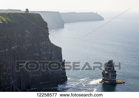 Stock Photo - Large sea stack rock structure in the ocean with dramatic rock cliff shoreline and castle turret in the distance; Liscannor, County Clare, Ireland. Fotosearch