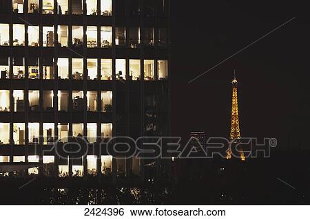 Stock Photograph - Lights illuminated in the offices of an office building at nighttime with an illuminated Eiffel tower in the background; Paris, France. Fotosearch