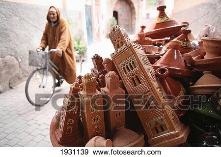 Stock Photo - Local man wearing long-hooded cloak, known as a djellaba, passing on his bicycle. Tajine pots and models of minarets of Koutoubia Mosque for sale at a street stall in the souk Marrakesh, Morocco, Africa., Marrakesh, Morocco, Africa.. Fotosearch