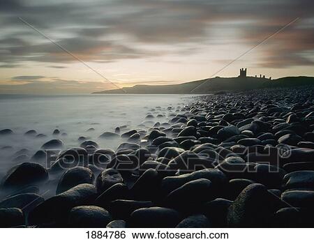 Stock Photograph - Looking Along Boulder Covered Beach Towards Dunstanburgh Castle At Dawn. Fotosearch