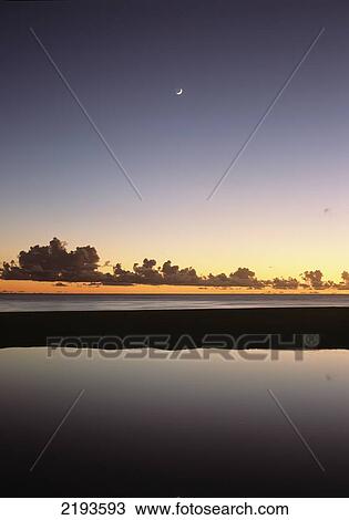 Stock Image - Looking over shallow pool at dusk to moon setting off West coast; Barbados. Fotosearch