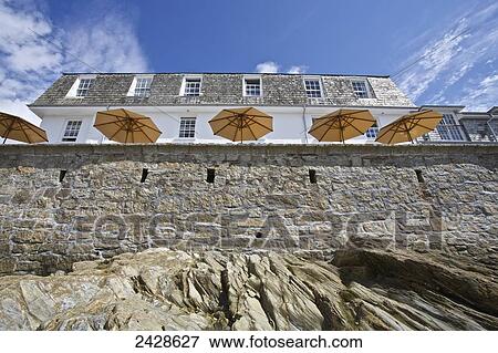 Stock Photo - Looking up a wall from the shore to a building and umbrellas on a patio on the seaside; St. Mawes, Cornwall, England. Fotosearch