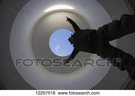 Stock Photograph - Low angle view of a person standing with arms raised and a round opening to blue sky above; Kielder, Northumberland, England. Fotosearch