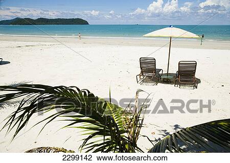Stock Image - Malaysia, Pulau Langkawi, Deck chairs under parasol on white sandy beach with palm trees overlooking blue sea; Pantai Cenang (Cenang beach). Fotosearch