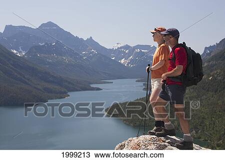 Male And Female Hiker Standing On A Rock Cliff Looking Out Onto A Lake With Mountains And Blue Sky; Waterton Alberta Canada View Large Photo Image Stock Image - Male And Female Hiker Standing On A Rock Cliff Looking Out Onto A Lake With Mountains And Blue Sky; Waterton Alberta Canada. Fotosearch