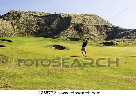 Stock Image - Male golfer on fairway chipping to the green framed by grassy hills with blue sky; County Clare, Ireland. Fotosearch