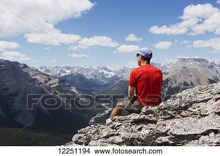 Picture - Male hiker sitting on a rocky cliff ledge overlooking a river valley below with mountain range in the distance with blue sky and clouds; Alberta, Canada. Fotosearch