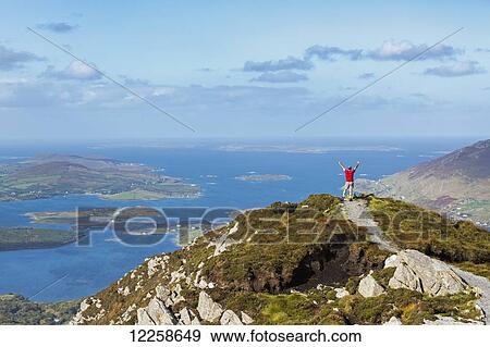 Stock Photo - Male hiker with arms in the air on top of mountain overlooking the ocean with islands in the distance; Letterfrack, County Galway, Ireland. Fotosearch