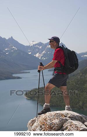 Stock Image - Male Hiker With Backpack And Hiking Poles Standing On A Rock Cliff Looking Out Onto A Lake With Mountains And Blue Sky; Waterton Alberta Canada. Fotosearch