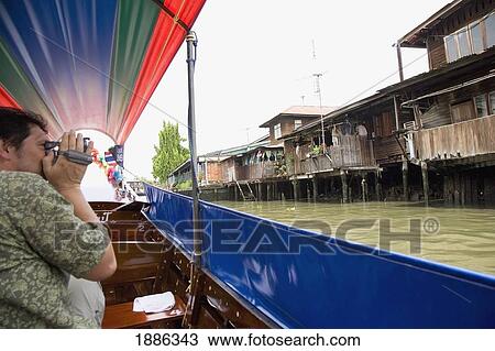 Stock Image - Male Tourist Videoing Houses From Longtail Boat On Klong Bangkok Noi. Fotosearch