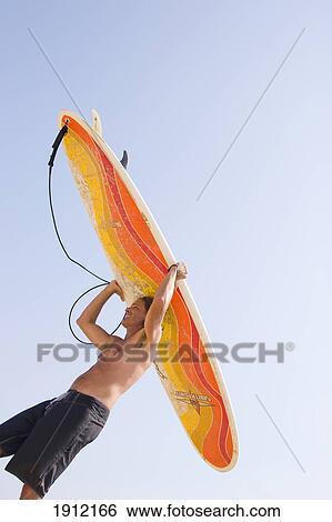 Stock Photograph - Man Carrying Surfboard, Low Angle View Against Blue Sky, Ditch Plains, Montauk, Long Island, New York, Usa. Fotosearch