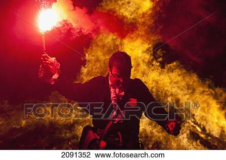 Stock Image - Man Dressed As Pirate From Southover Bonfire Society In Front Of Large Bonfire Holding Red Flare, Bonfire Night, Lewes, East Sussex, Uk. Fotosearch