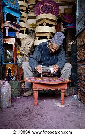 Picture - Man hand painting a table with others piled up in the souk; Marrakesh, Morocco. Fotosearch