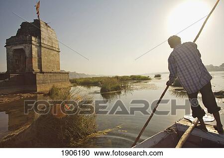 Stock Image - Man In Boat Navigating On Narmada River Near Temple Ruins, Rear View, Maheshwar, Madhya Pradesh, India. Fotosearch