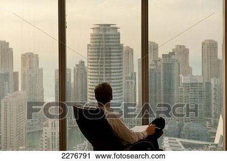Stock Image - Man in office looking out from large windows to office and residential tower blocks in Dubai Marina; Dubai, United Arab Emirates. Fotosearch