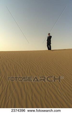 Stock Photograph - Man in smart suit making phone call on top of sand dune at dusk; Dubai, United Arab Emirates. Fotosearch