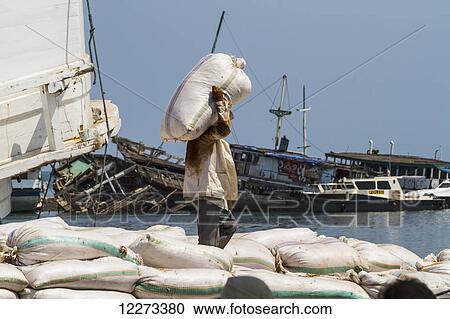 Stock Image - Man loading a sack of cargo on a Pinisi, a traditional two masted wooden cargo sailing ship moored in Paotere Harbour, Makassar (Ujung Pandang), South Sulawesi, Indonesia. Fotosearch