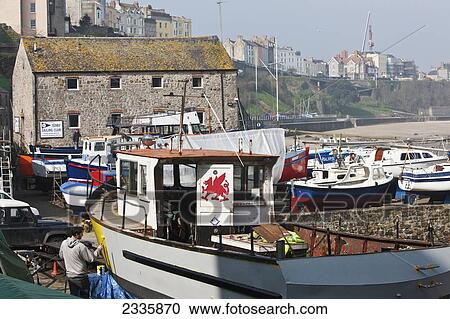 Stock Image - Man Painting Boat, Tenby Harbor With North Beach In Background; Pembrokeshire Coast Path, Wales, United Kingdom. Fotosearch
