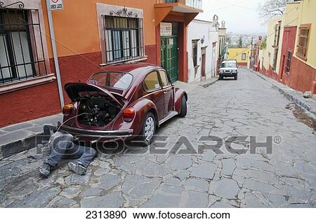 Stock Image - Man repairing volkswagen beetle parked on cobblestone street; San Miguel de Allende, Guanajuato, Mexico. Fotosearch