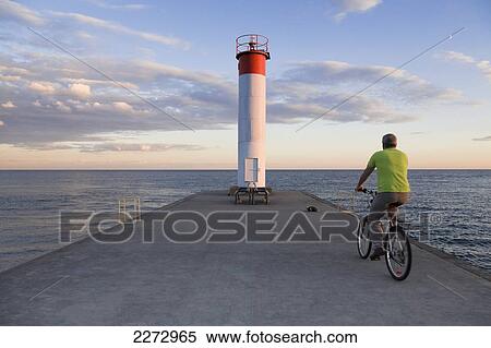 Stock Photography - Man riding a bicycle toward a lighthouse at sunset by lake ontario; whitby ontario canada. Fotosearch