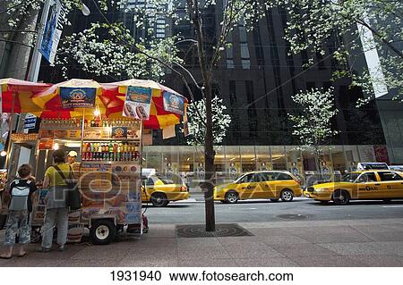 Stock Image - Man Selling Hot Dogs And Taxis Passing In Front Of The Museum Of Modern Art Also Known As Moma, Manhattan, New York, Usa. Fotosearch