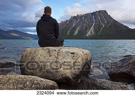 Picture - Man sitting on a rock by Kusawa Lake with Ark Mountain in the background at dusk; Yukon, Canada. Fotosearch