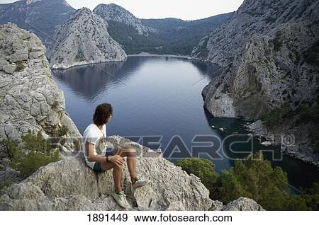 Stock Photo - Man Sitting On Cliff, Looking At View. Fotosearch