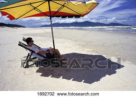 Stock Image - Man Sitting On Deckchair Under Umbrella On Beach, Side View. Fotosearch