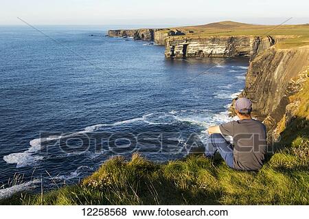Man sitting on grassy cliff edge overlooking the dramatic rock cliffs along the shoreline at sunset; Kilkee, County Clare, Ireland View Large Photo Image Stock Photo - Man sitting on grassy cliff edge overlooking the dramatic rock cliffs along the shoreline at sunset; Kilkee, County Clare, Ireland. Fotosearch
