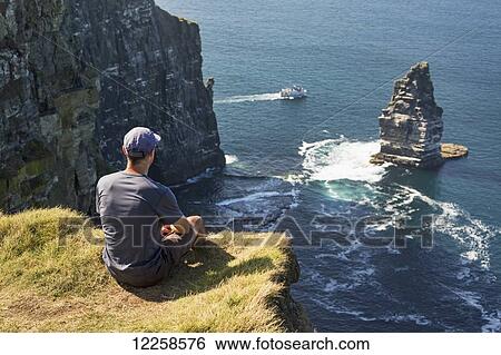 Stock Photograph - Man sitting on grassy cliff's edge overlooking large sea stack rock structure in the ocean with tour boat; Liscannor, County Clare, Ireland. Fotosearch